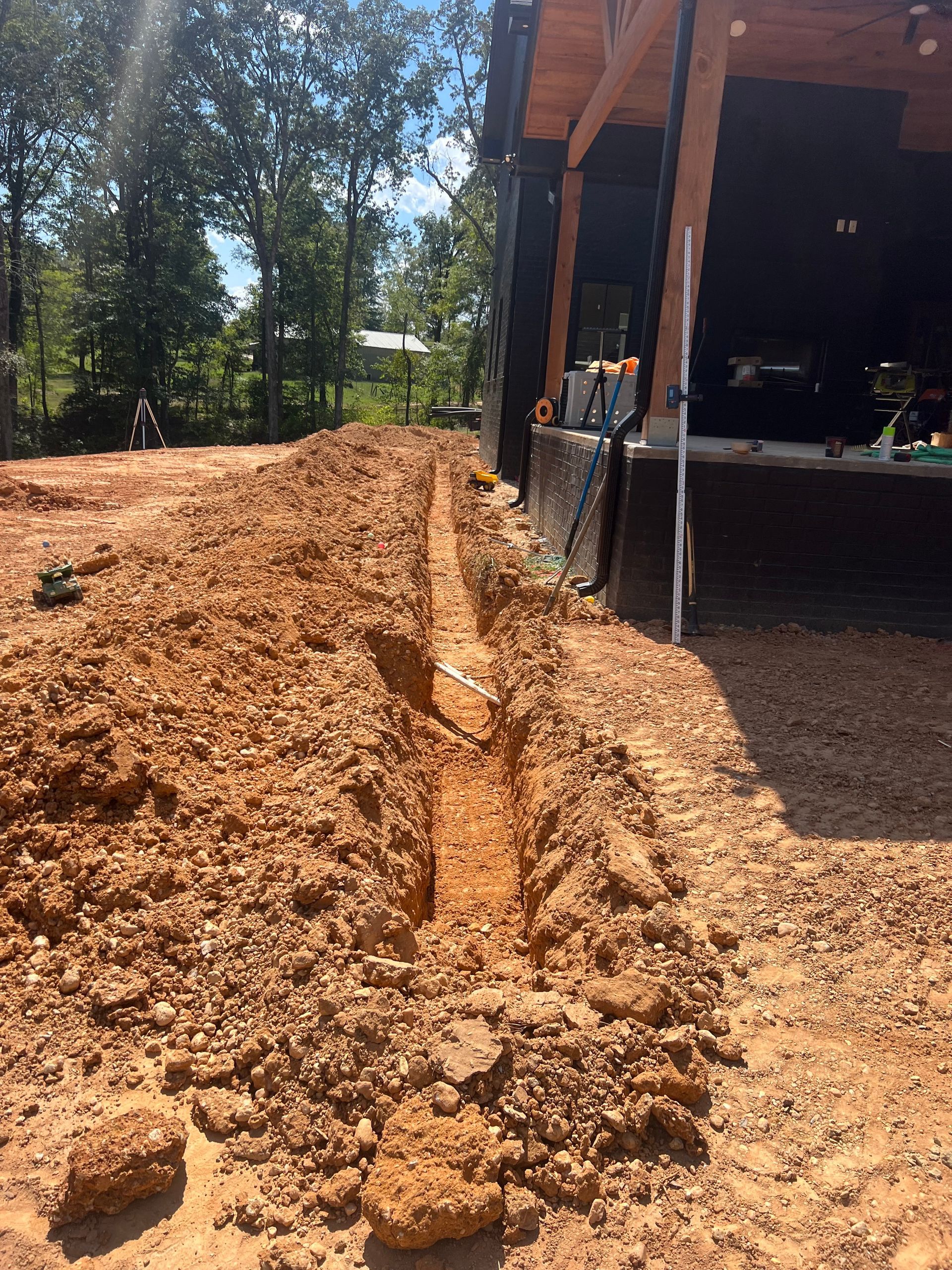 Freshly dug trench beside a house under construction, with piles of reddish soil and trees in the background