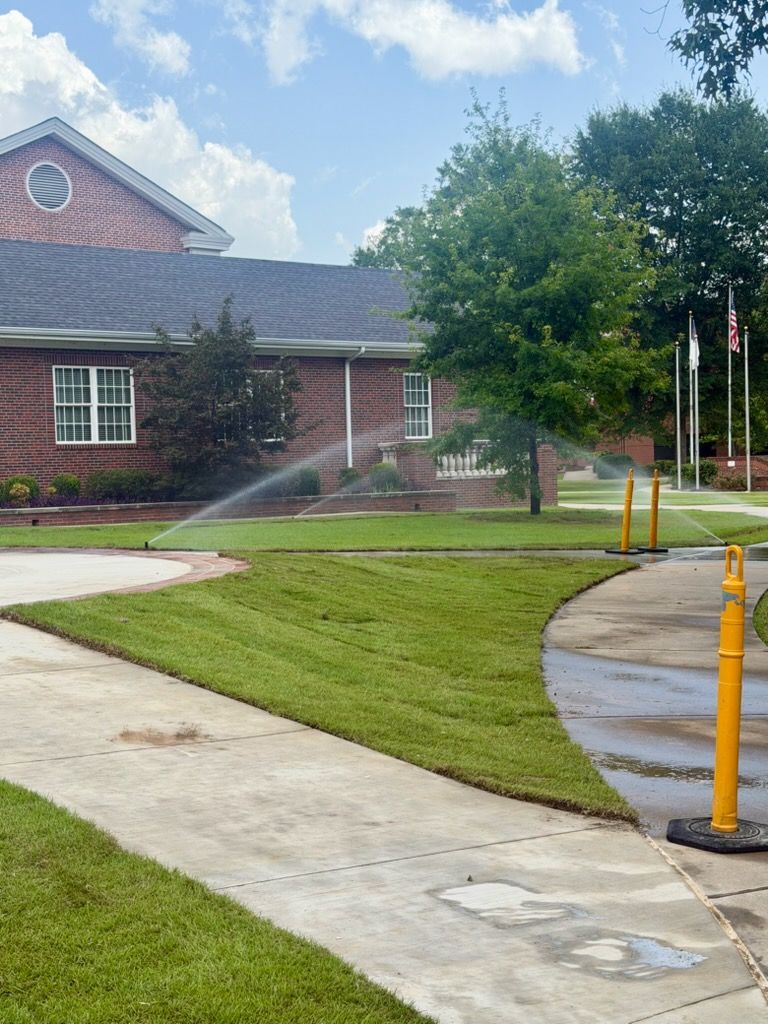 Sprinklers watering a lawn beside a brick building with curved sidewalk and yellow bollards