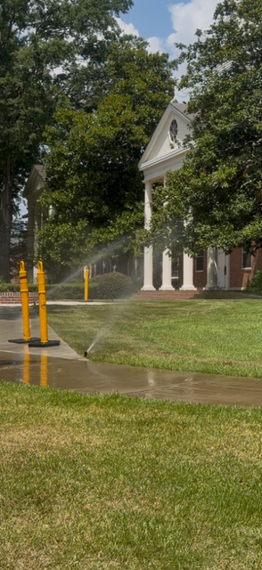 Flooded campus walkway beside a white columned building, with yellow bollards and trees after rain.