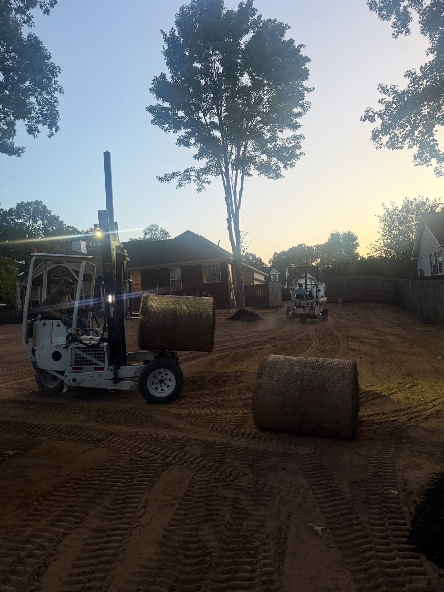 Construction site with two small machines and rolled hay bales on a dirt lot at sunrise.