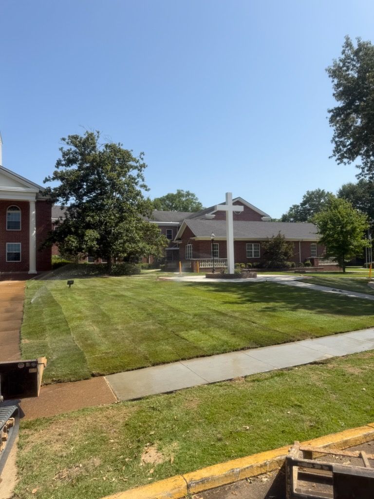 Campus lawn with a white cross monument, brick buildings, and trees under a clear blue sky