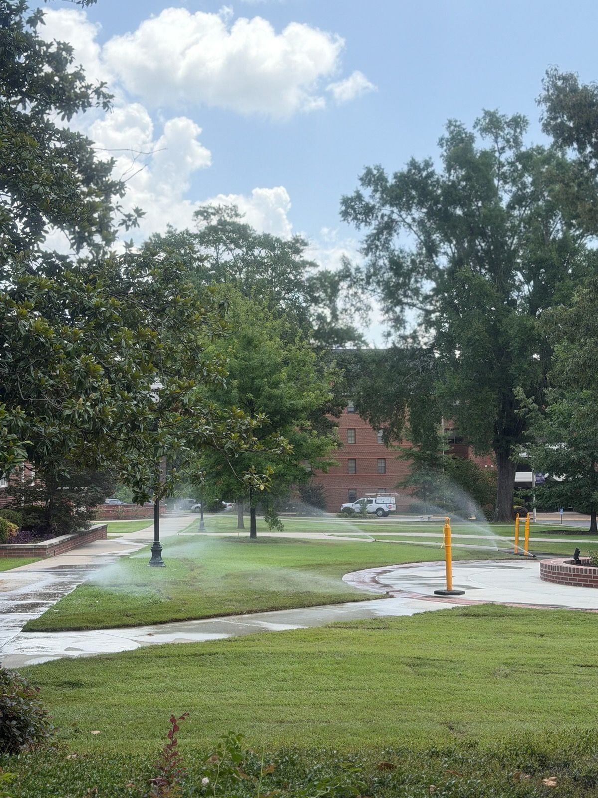 Rain-soaked park path and lawn with trees, a red brick building in the background, and a yellow post.