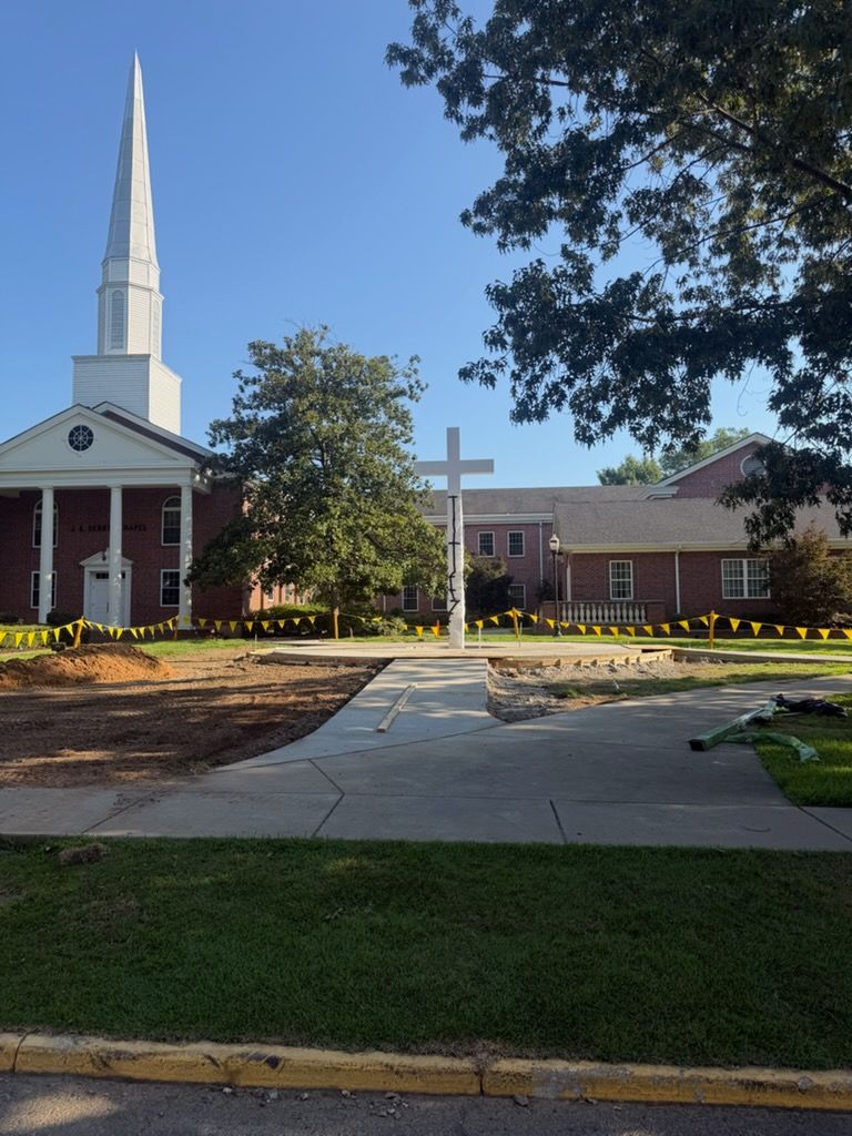 Church with tall white steeple, white cross in front, and brick buildings along a walkway under a blue sky.
