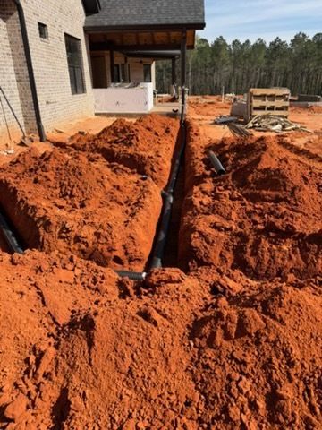 Red dirt trench beside a house under construction, with a black pipe running along the excavation.