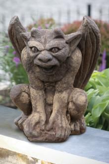 A gargoyle statue is sitting on a ledge in a garden.