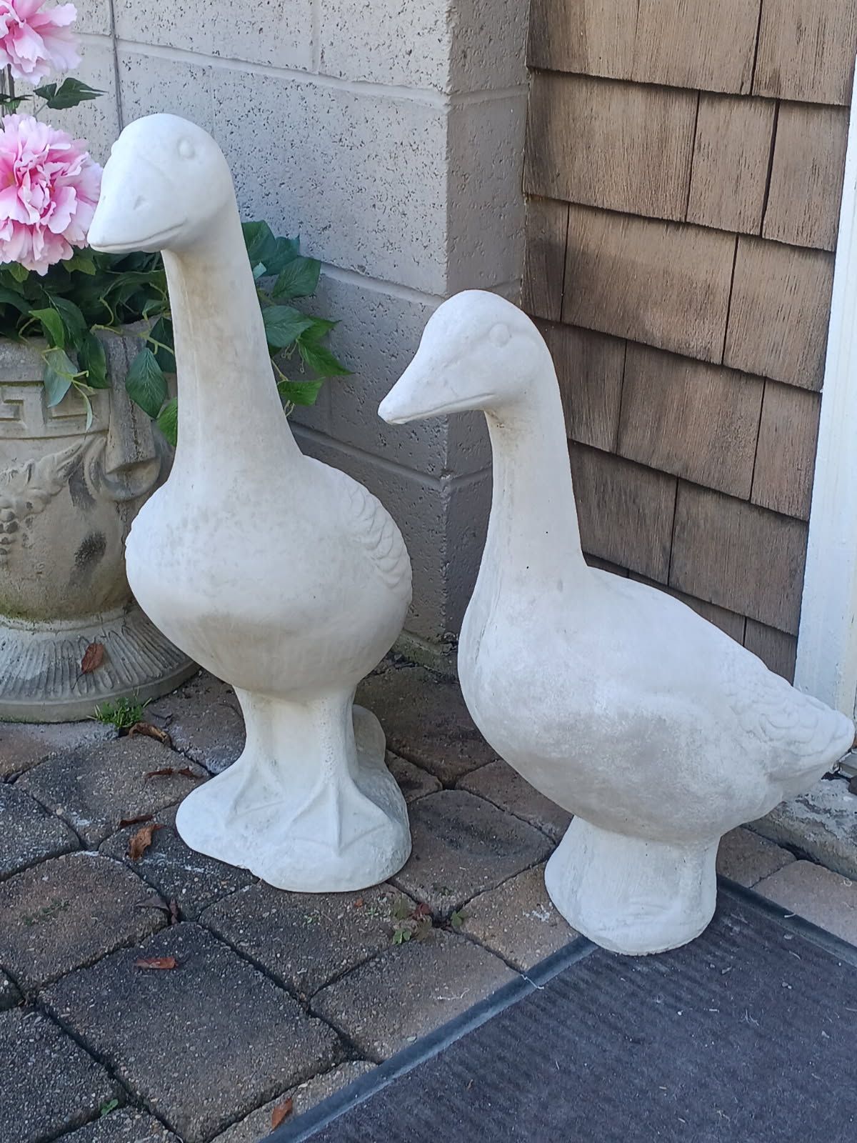 Two white goose lawn statues on a stone patio beside a wall and flowers