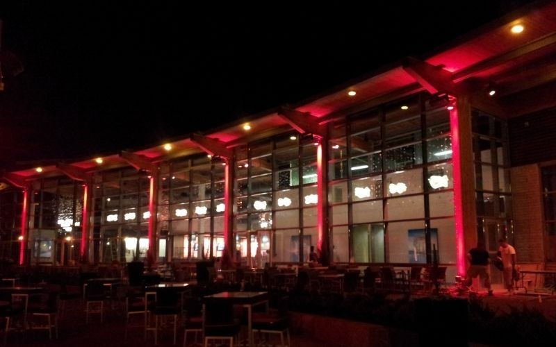 Night view of a building with red lighting on the pillars, outdoor seating area, and dark sky.