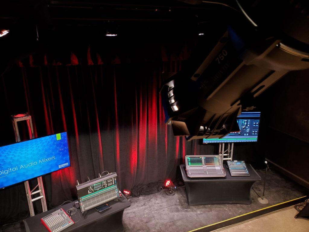 A room with a red curtain , a table , a monitor , and a projector.