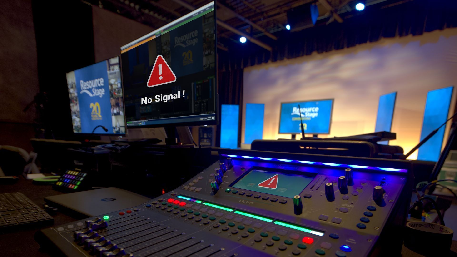 Audio mixing console in a dark studio with monitors showing a “No Signal” warning on stage