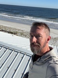 A man with a beard is standing on a roof overlooking the ocean.