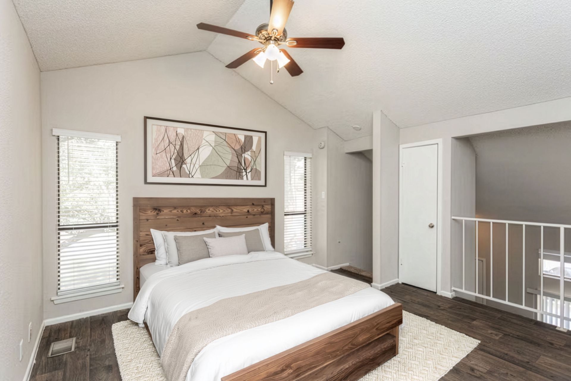 Bright bedroom with a wood bed, ceiling fan, white walls, windows, and a balcony railing beyond the doorway