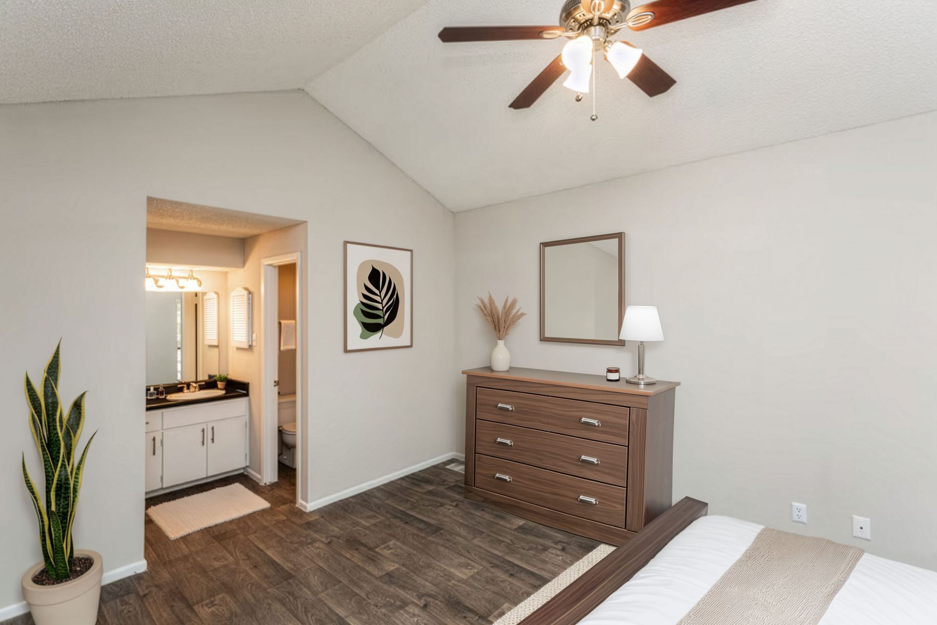 Bright bedroom with wood dresser, ceiling fan, potted plant, and bathroom doorway.