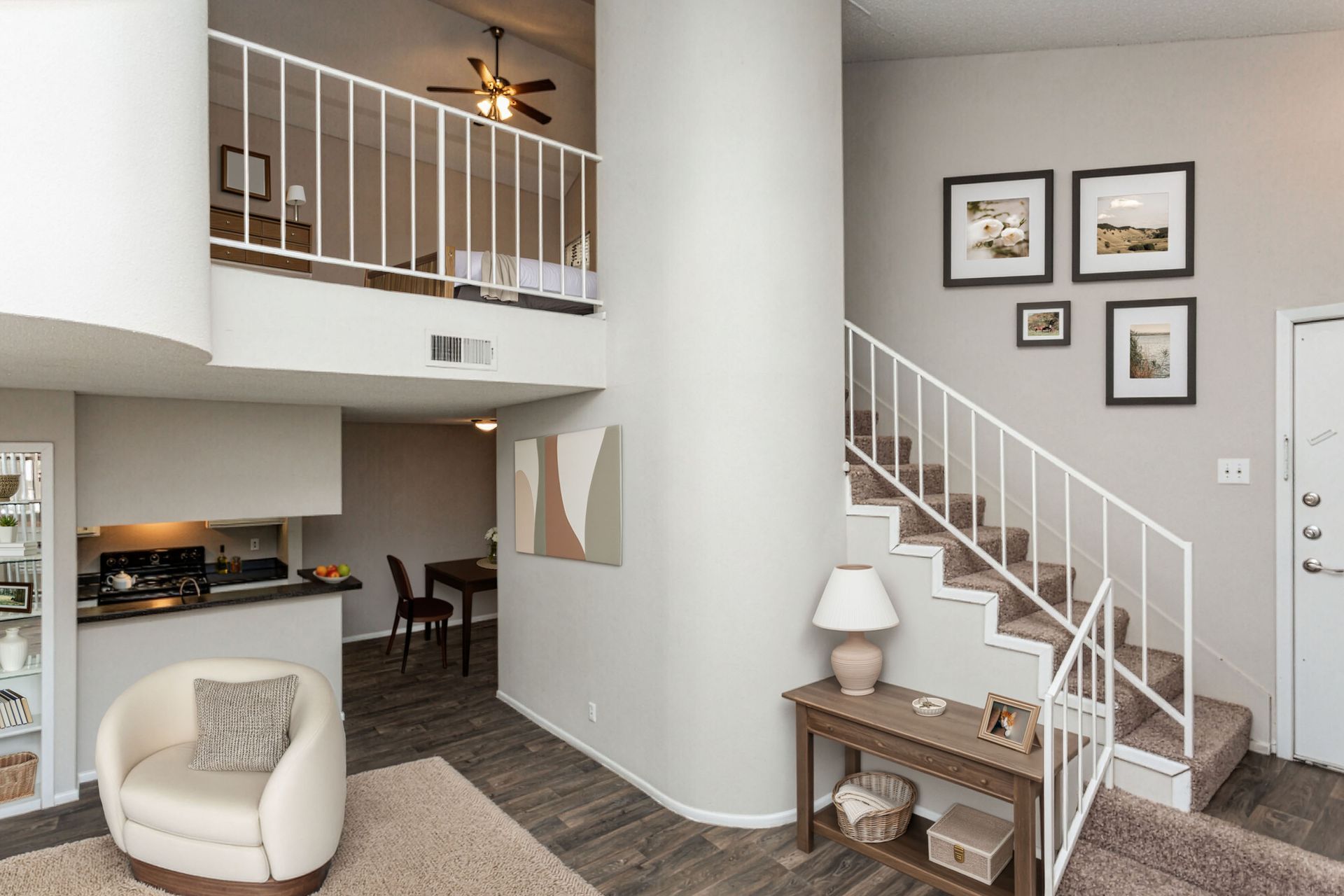 Bright open living room with staircase, loft railing, neutral chair, and small console table with lamp and framed art.