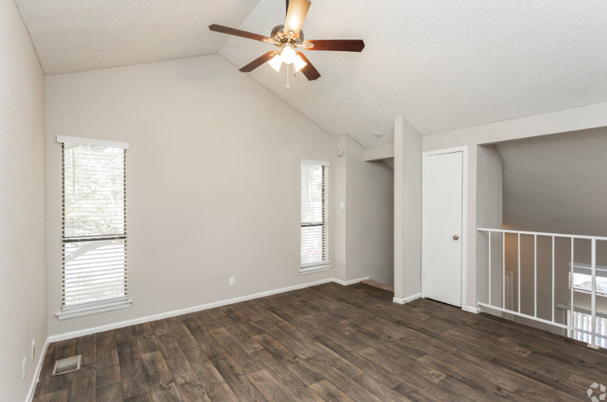 Empty beige bedroom with dark wood floors, ceiling fan, and a view of the upstairs railing.