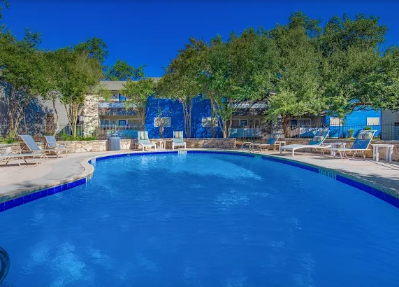 Blue resort pool surrounded by lounge chairs and trees under a clear sky