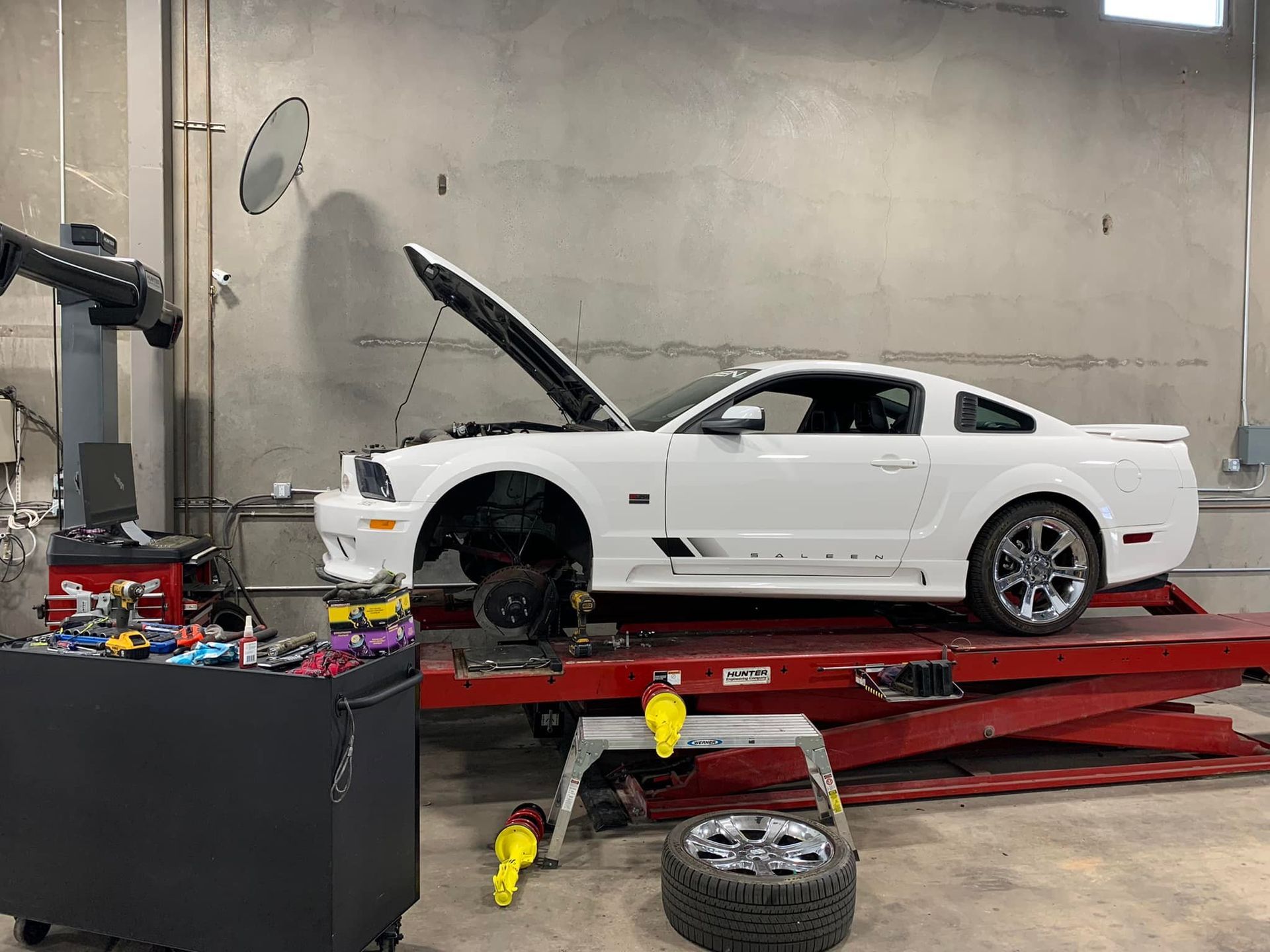 White Mustang on a car lift in a garage with its hood open and a tire removed.