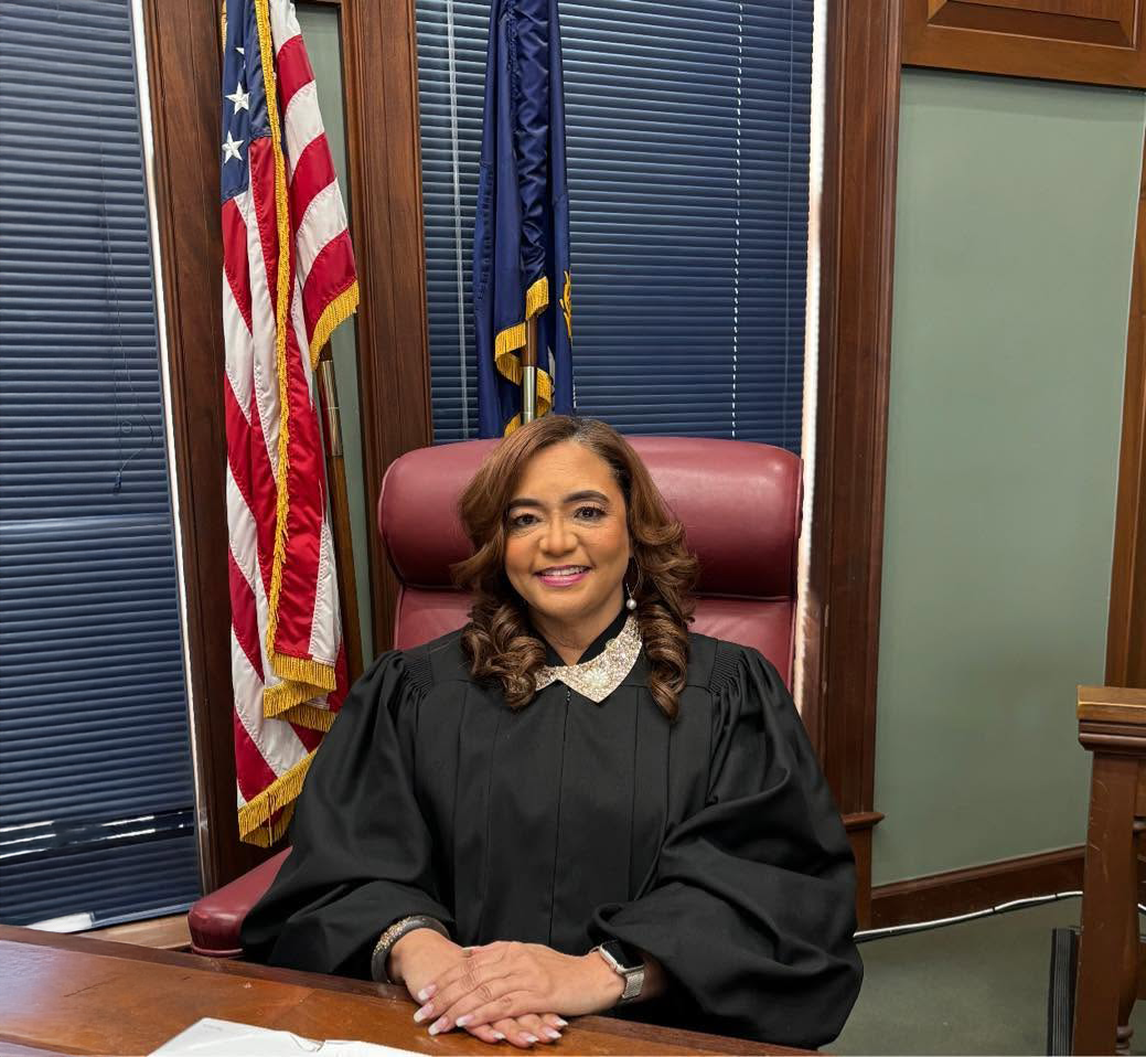 Judge seated behind a desk in a courtroom, smiling, with U.S. and state flags behind her.