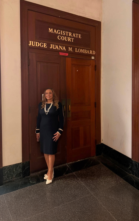 Woman in black dress standing by a wooden door labeled “Magistrates Court” and “Judge Joan A. …”