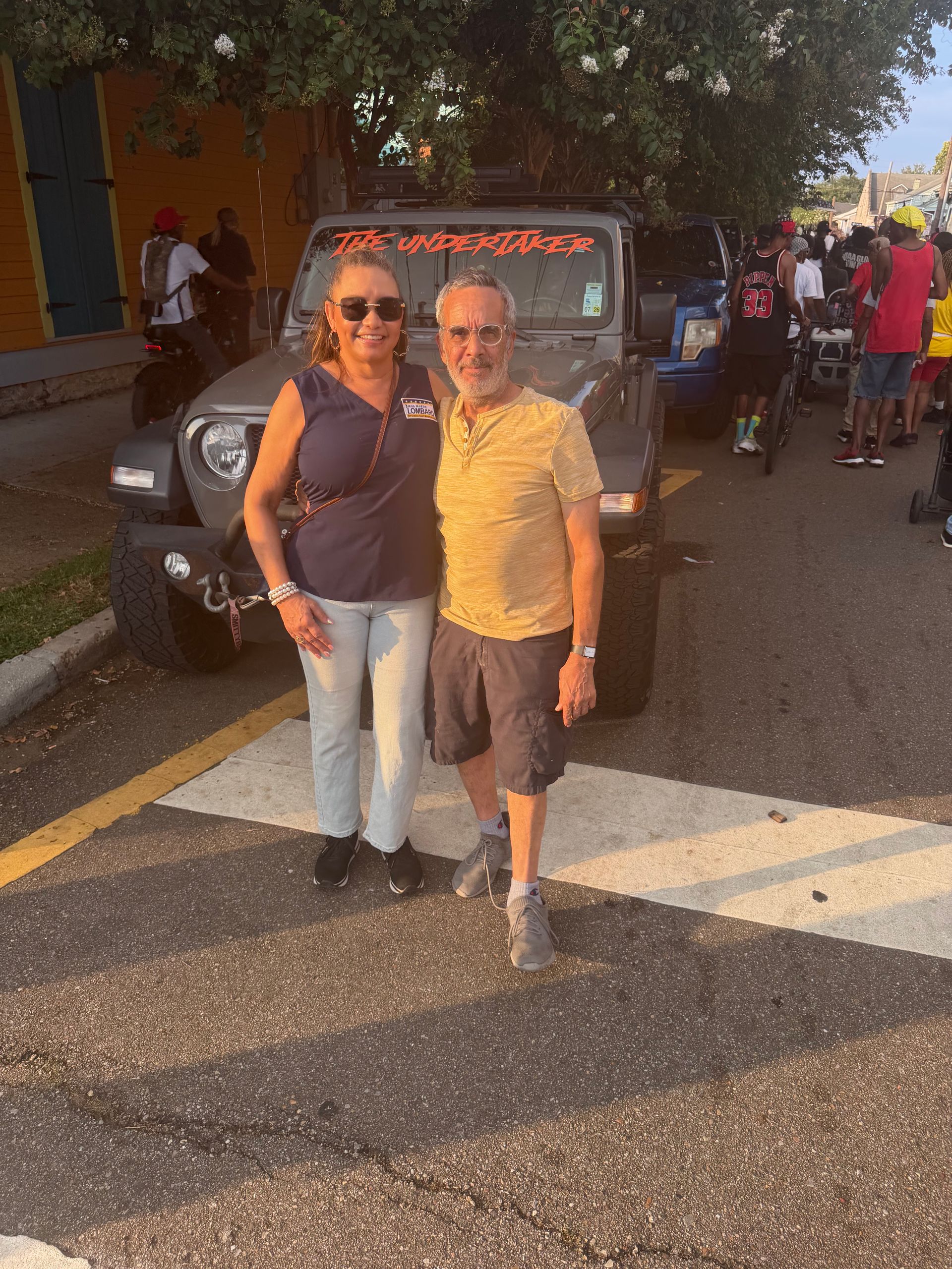 Two people posing in front of a Jeep on a street at sunset, smiling and wearing sunglasses.