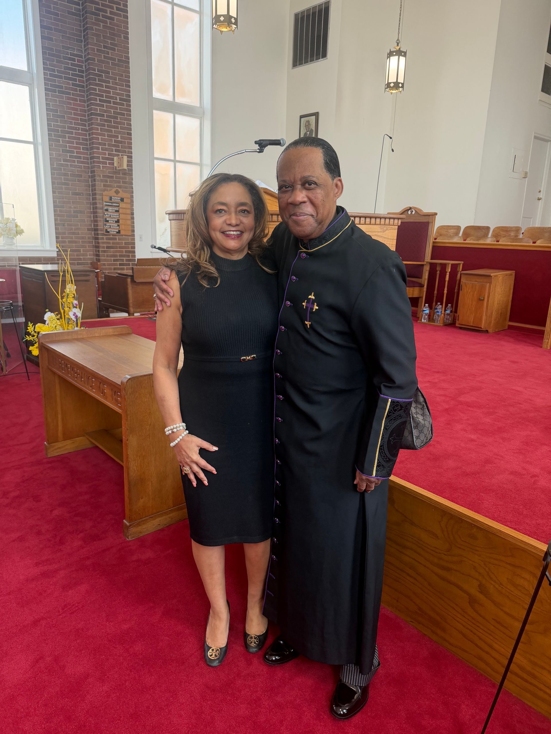 Two people standing together in a church sanctuary with red carpet and wooden pews.