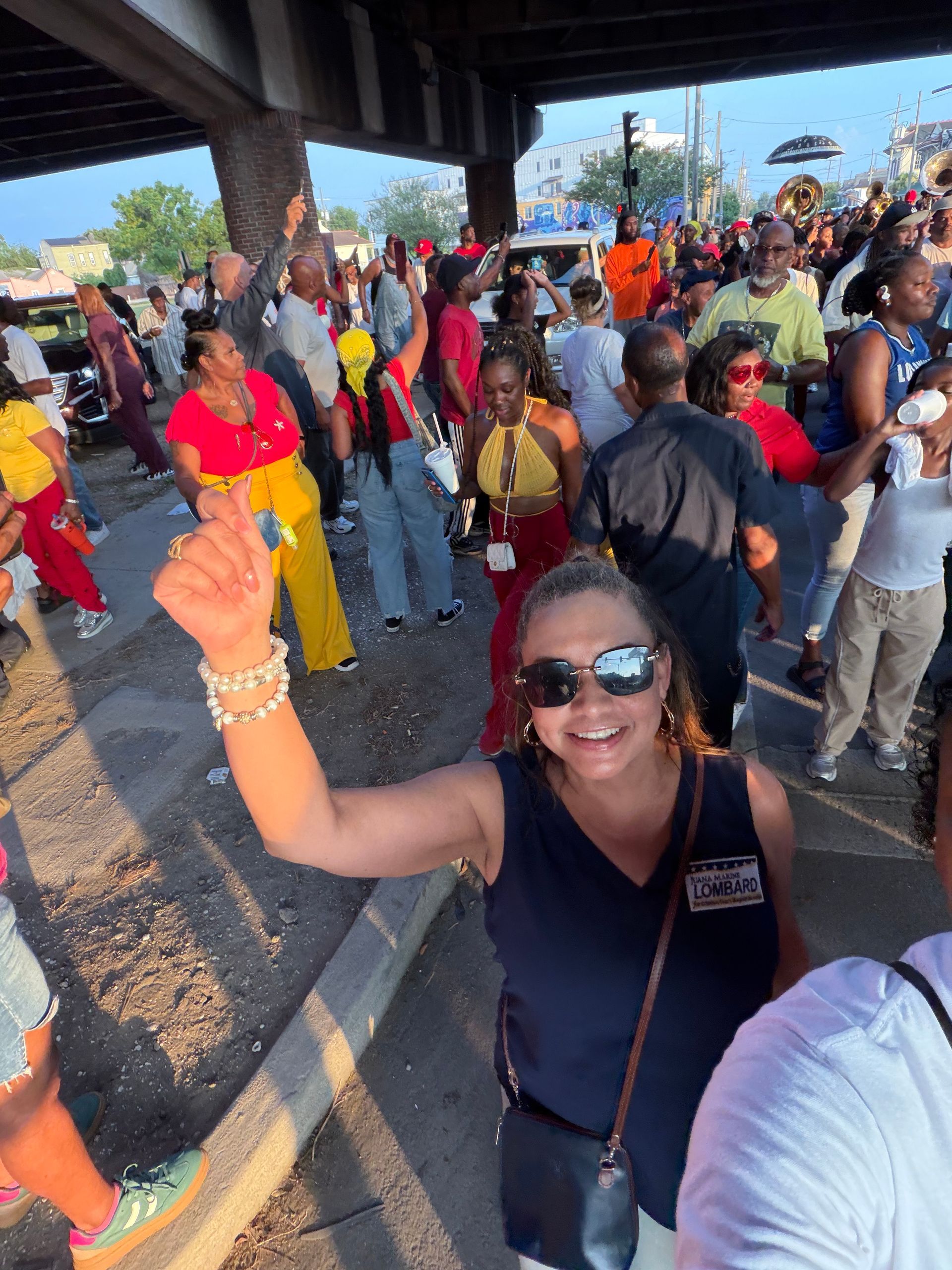 Crowd dancing and cheering at an outdoor event, with a woman in sunglasses raising her arm in front.