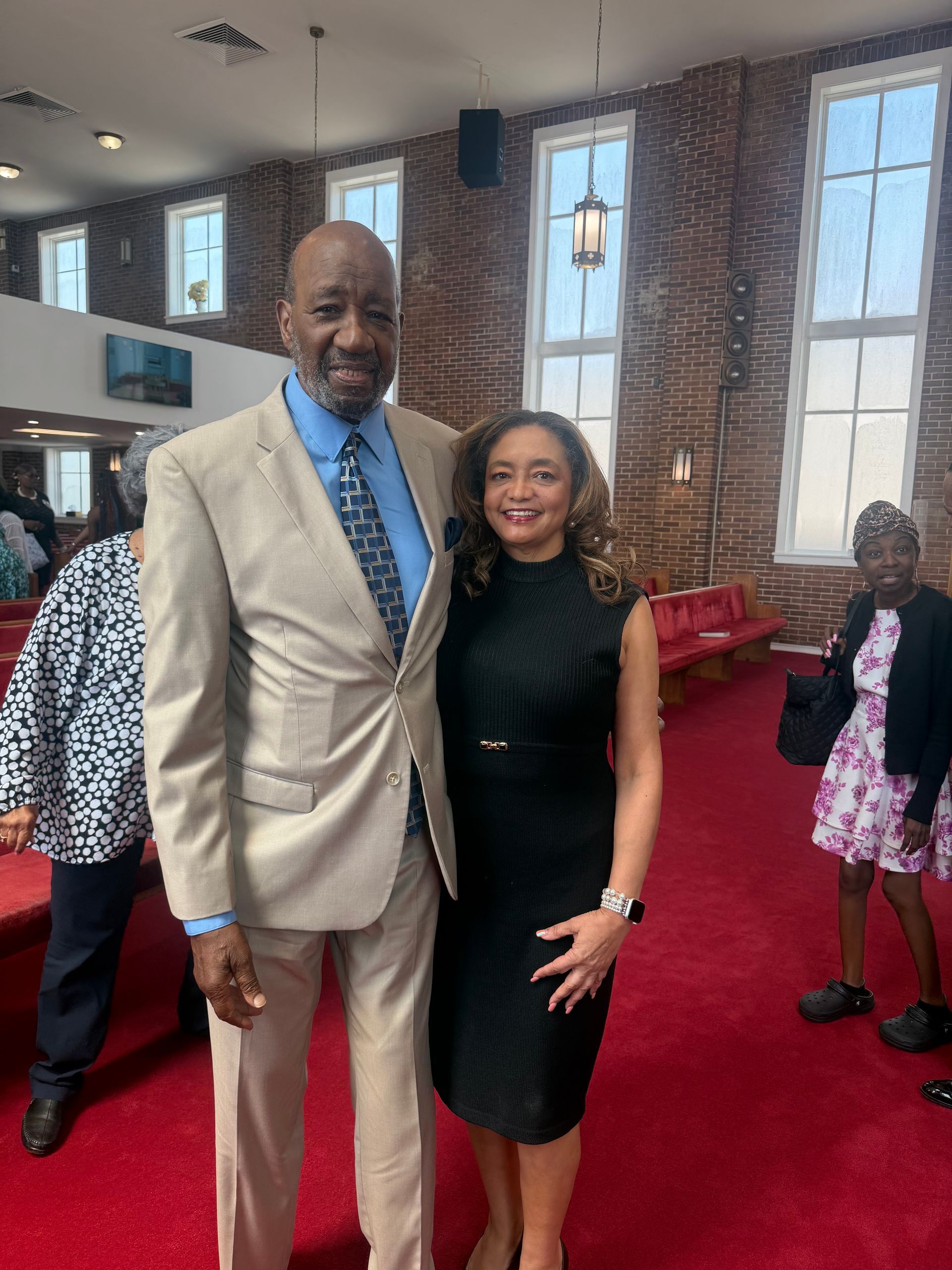 Two people posing on a red-carpeted church interior, smiling beside brick walls and tall windows.