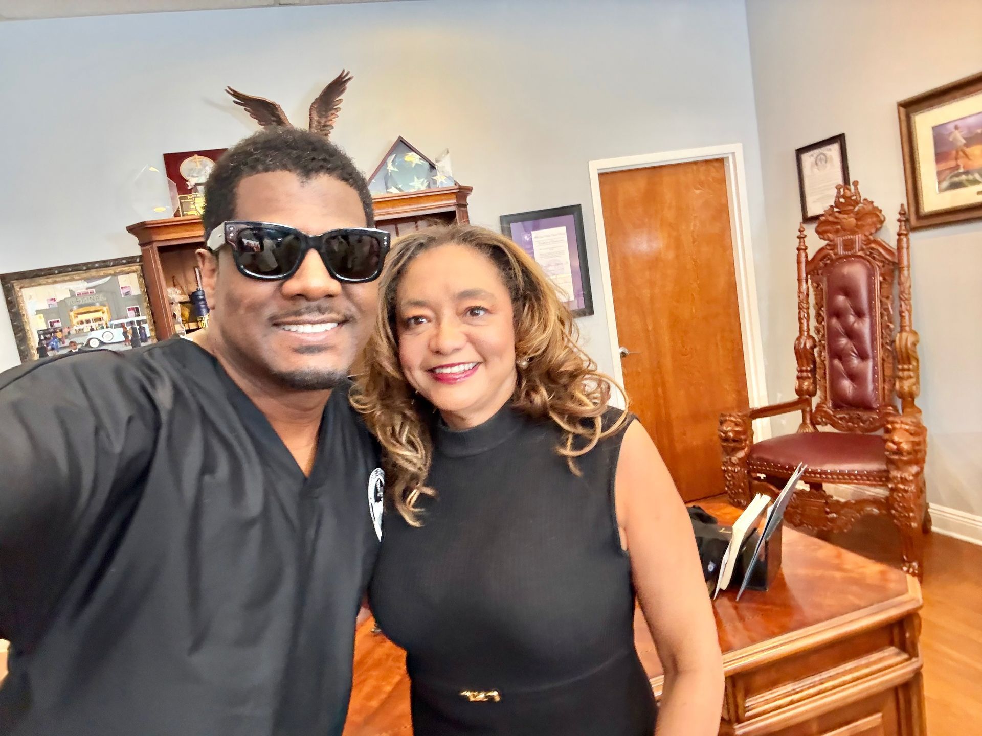 Two people smiling in an office, standing beside a wooden desk and ornate chair.