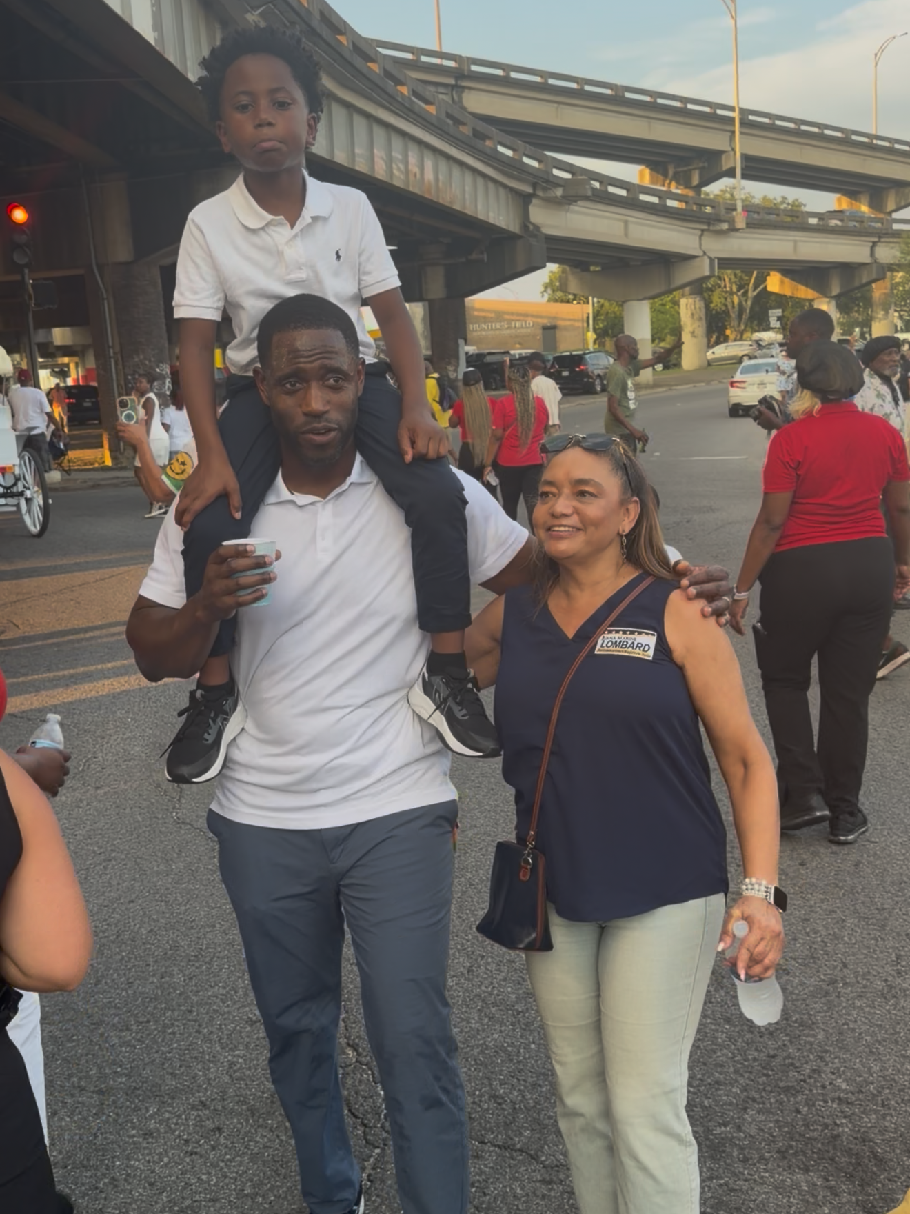 Man carrying a child on his shoulders beside a smiling woman on a busy street under an overpass