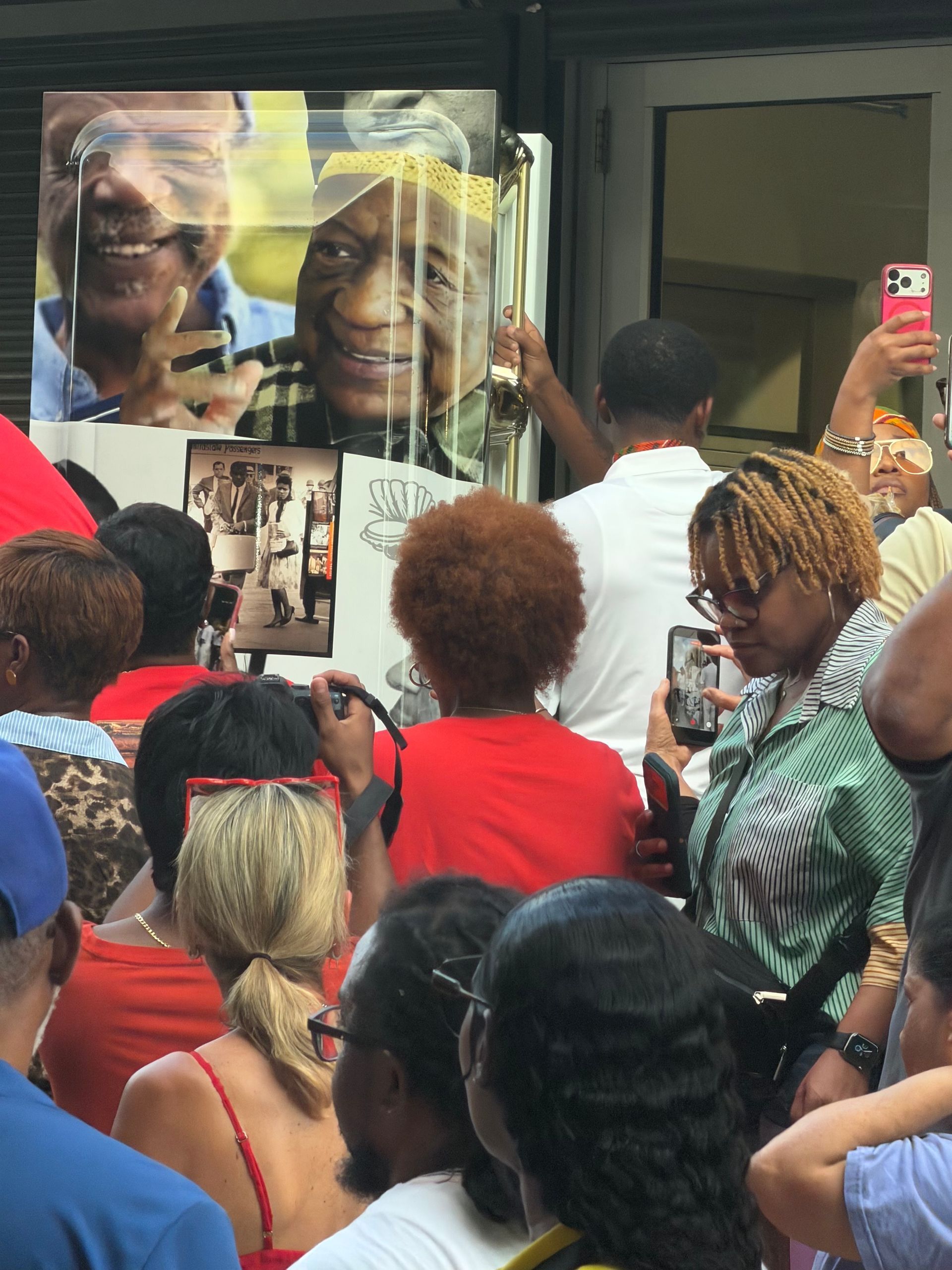 Crowd gathered around a storefront display of portraits, with people taking photos on their phones.
