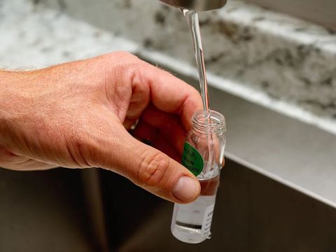 A hand holds a small glass vial under a running faucet to collect a water sample.