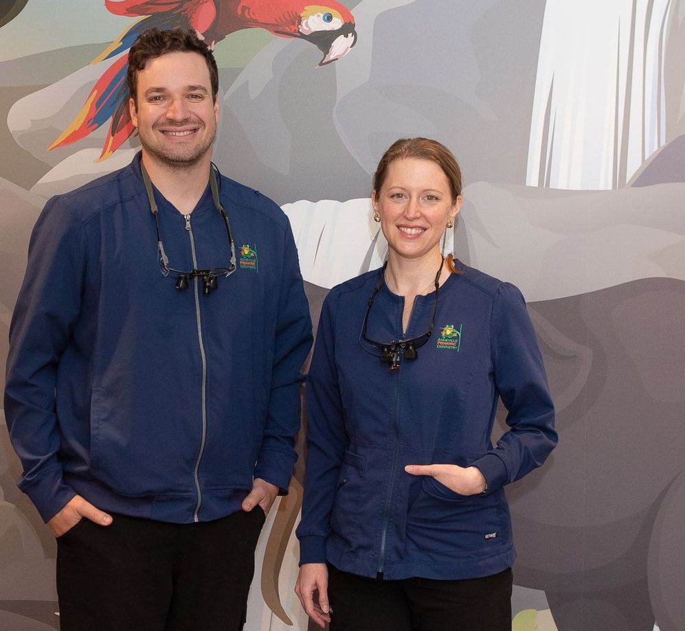 A man and a woman are posing for a picture in front of a painting of a parrot Asheville Pediatric Dentistry (828) 277-6788