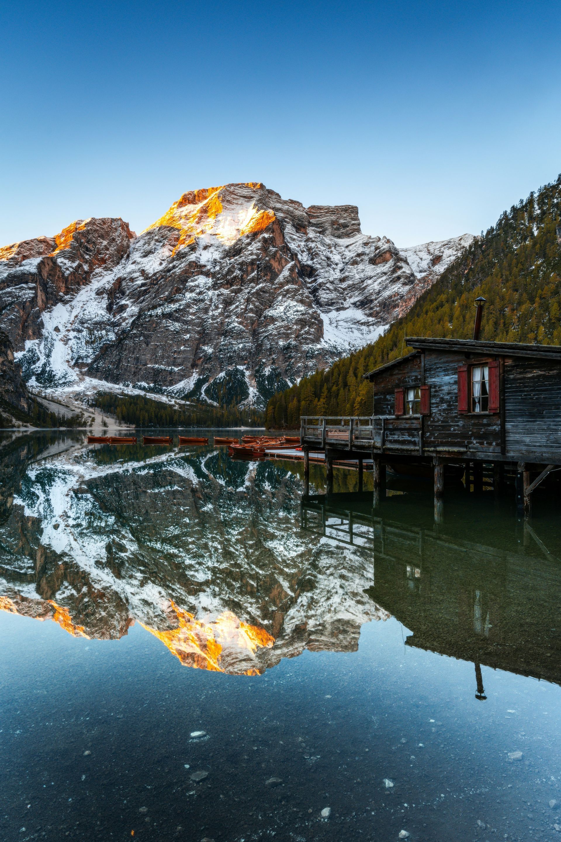 Snowy mountain reflecting in a still lake; wooden cabin on a dock, blue sky.