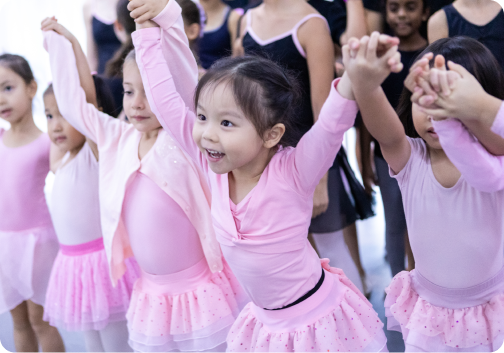 A group of young girls in pink tutus are holding hands in a dance studio.