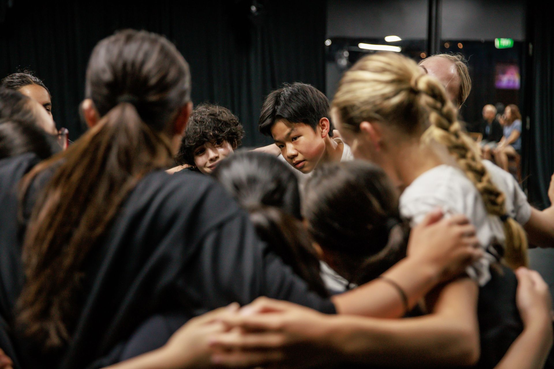 A group of people standing in a close circle, arms around each other's shoulders, inside a dimly lit performance space.