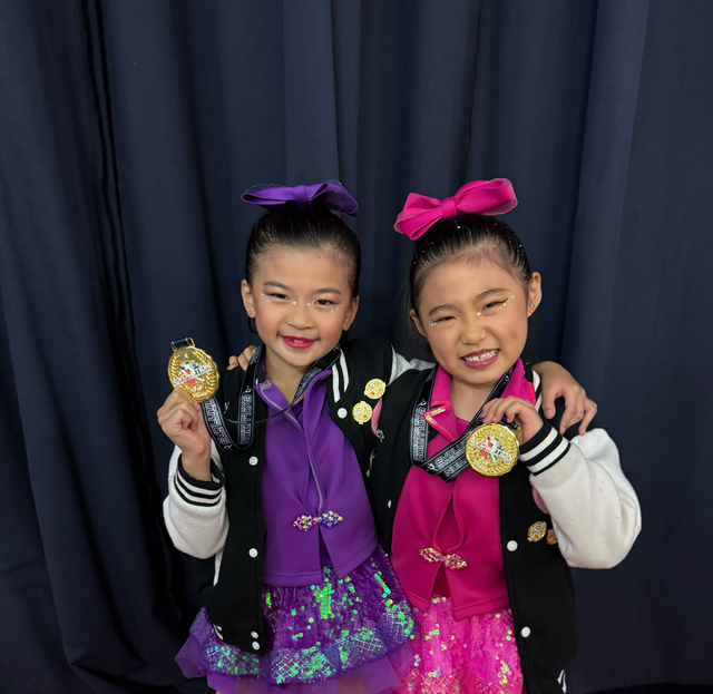 Two children wearing matching black varsity jackets and colorful hair bows hold gold medals while smiling against a curtain.