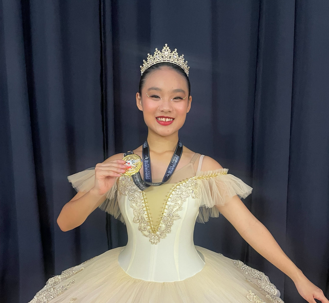 A smiling ballerina wearing a crown and a cream-colored tutu holds a medal against a dark blue curtain backdrop.