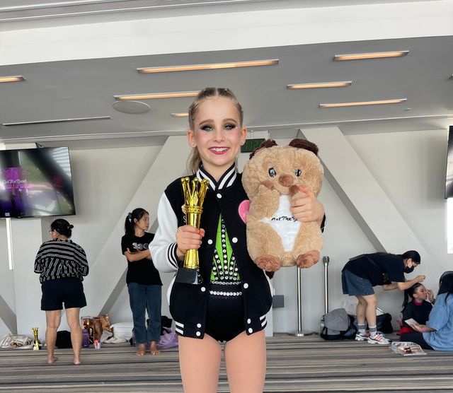 A young person wearing a varsity jacket holds a gold trophy and a plush toy in a brightly lit room with others nearby.