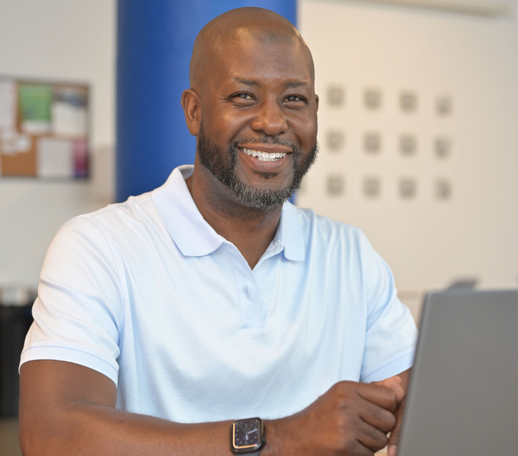 Man smiling, wearing a light blue polo shirt, looking at laptop. Indoors, neutral background.