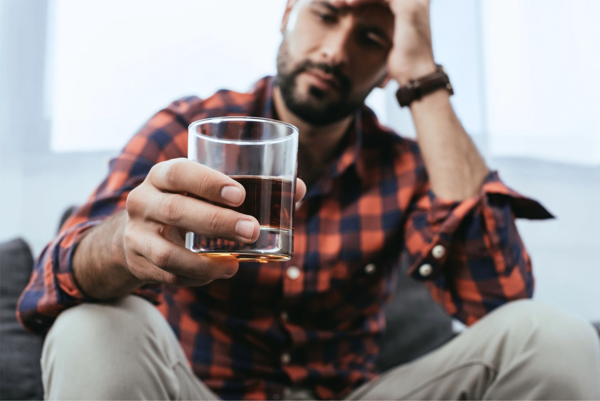 Man holds a glass of amber liquid, looking distressed while seated on a couch.