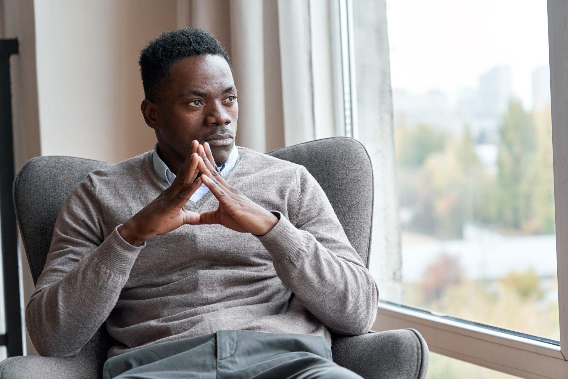 Man sitting in a chair, looking out a window. He has hands clasped, appearing contemplative.