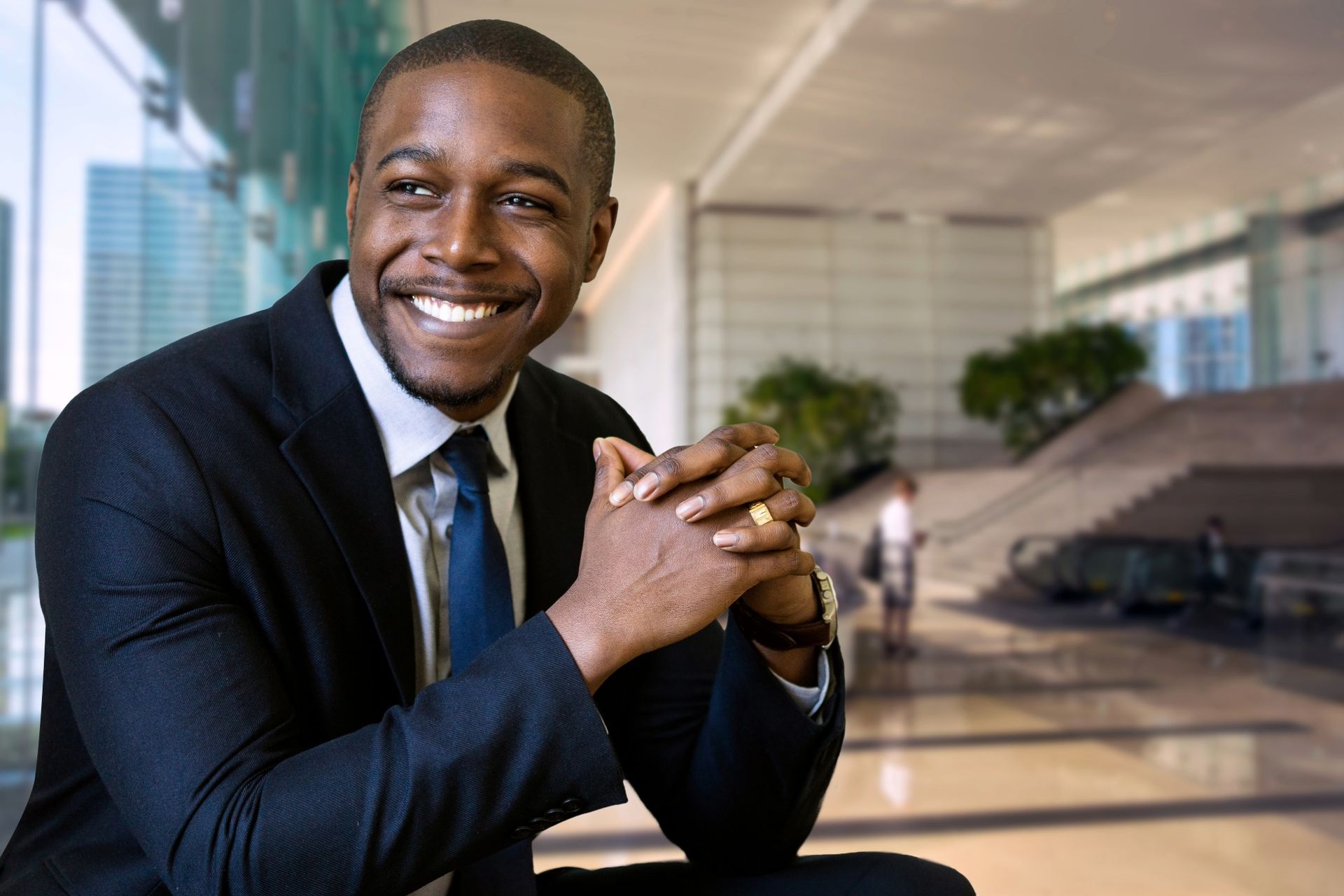 Man in a suit smiles, hands clasped, in a modern office.