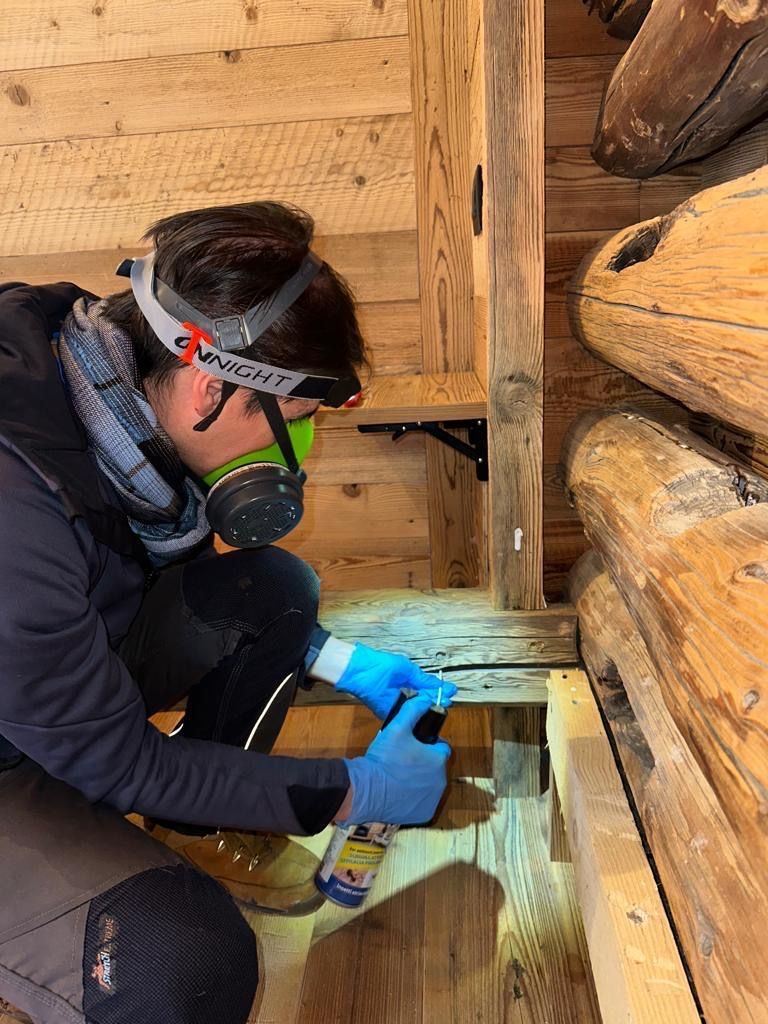 Una persona con i guanti blu usa un trapano all'interno di un'apertura nel muro di una baita di legno.