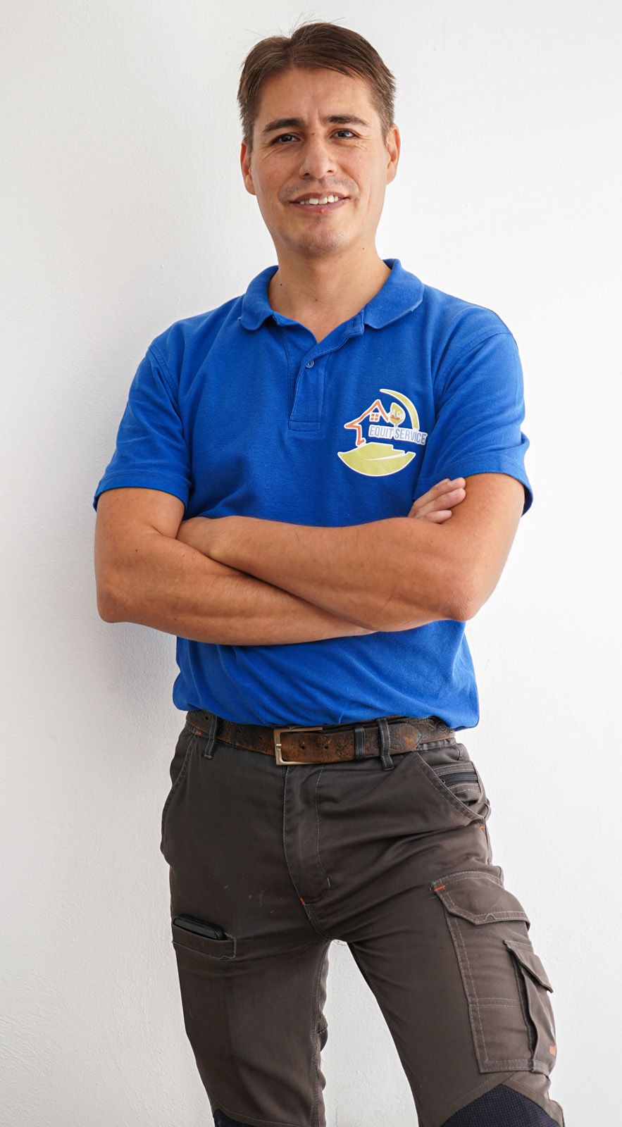 Persona con polo blu e pantaloni cargo grigi in piedi con le braccia incrociate su sfondo bianco.