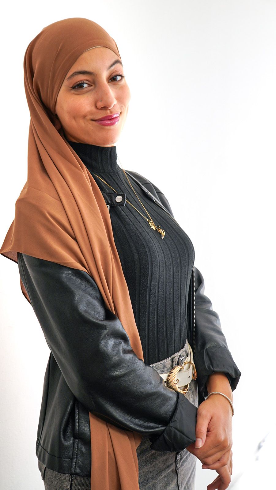 Donna con hijab marrone e giacca nera in posa su sfondo bianco.