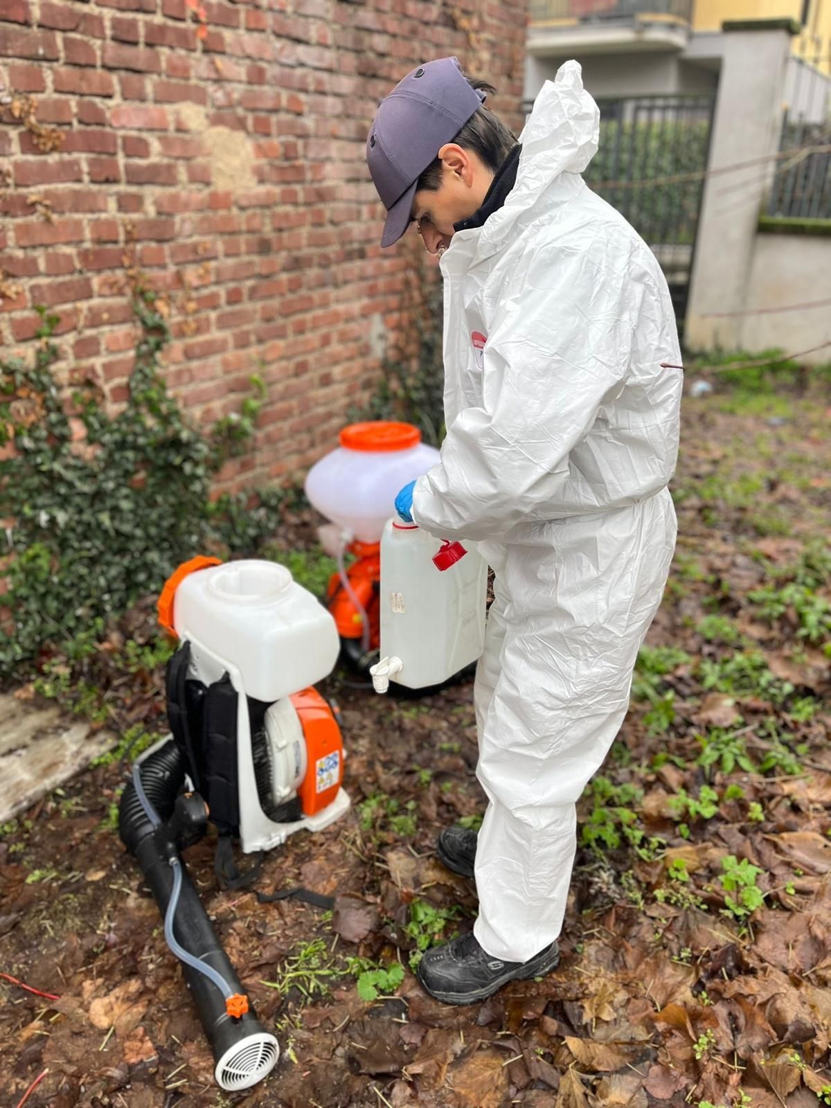 Un operaio con tuta protettiva bianca spruzza pesticida accanto a un muro di mattoni e a un giardino.