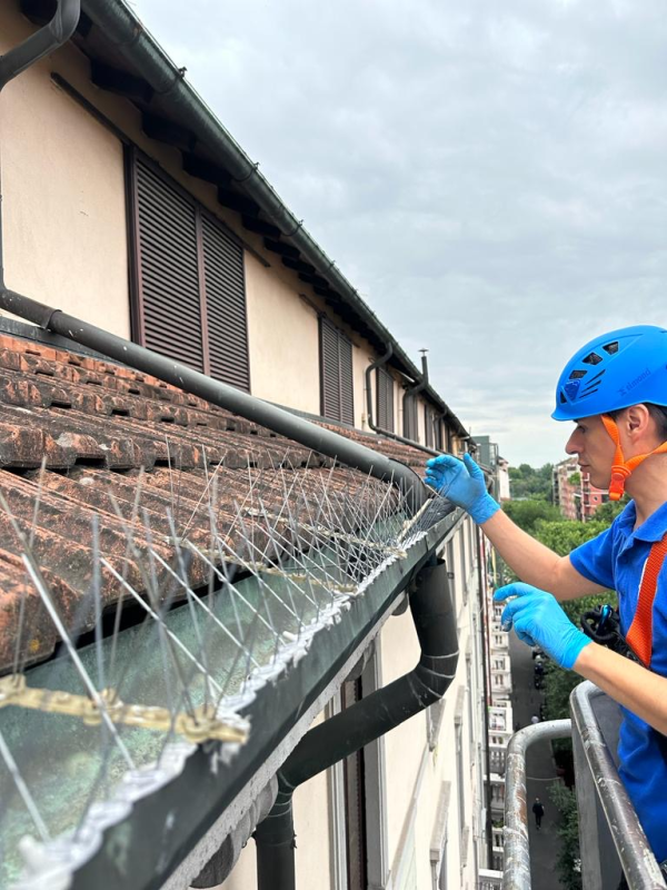 Un operaio con casco blu pulisce un tetto di tegole con imbracatura di sicurezza e rete protettiva in una giornata nuvolosa.
