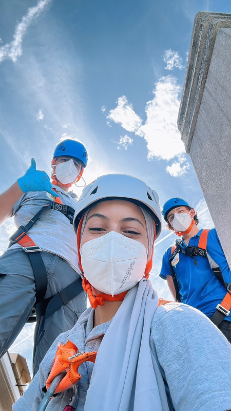 Tre alpinisti con casco, maschera e giacca posano su un tetto sotto un cielo azzurro.