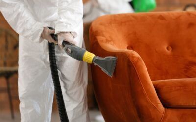 Una persona con indosso una tuta protettiva pulisce a vapore un divano arancione utilizzando un attrezzo manuale per tappezzeria.
