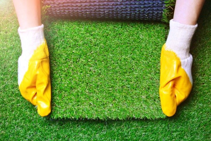 Hands wearing yellow work gloves laying down a fresh piece of artificial grass turf.