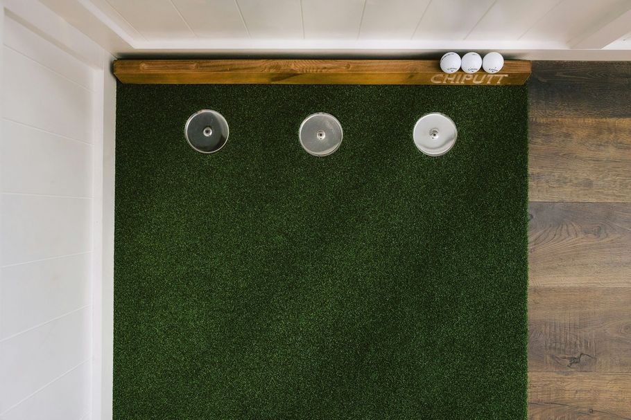 Top-down view of an indoor putting mat with three holes, a wooden frame, and three golf balls resting on the edge.